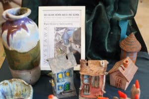 Ekspozycja ceramicznych domków i naczyń na stole przykrytym ciemnozielonym i granatowym materiałem. W centrum stoi biała ramka z plakatem informującym o wystawie prac wykonanych pod kierunkiem Pani Bożeny Sadowskiej. Na pierwszym planie widać kolorowe figurki domków z detalami takimi jak okna i kominy oraz wysoki wazon o szkliwieniu w odcieniach brązu, zieleni i fioletu