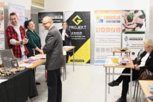 Stoiska organizacji. Przy stolikach z ulotkami stoją uśmiechnięte osoby rozmawiające ze sobą. W tle widoczne roll-upy z logotypami m.in. Projekt G, Fundacja 5 Horyzontów oraz Stowarzyszenie Modrzew.