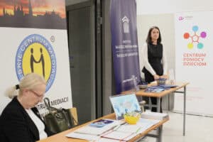 Stoiska informacyjne podczas wydarzenia. Na pierwszym planie kobieta siedząca przy stoliku Lubelskiego Centrum Mediacji. W tle kobieta stojąca przy roll-upie Instytutu na rzecz Państwa Prawa oraz Centrum Pomocy Dzieciom.