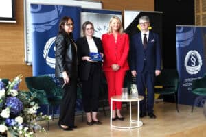 Cztery osoby stoją na scenie. Młoda kobieta w okularach trzyma otwarte etui z pamiątkową statuetką. Obok niej stoi kobieta w koralowym garniturze oraz mężczyzna w ciemnym garniturze.