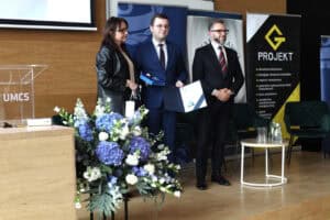 Uroczyste wręczenie nagrody. Mężczyzna w okularach i niebieskim garniturze trzyma dyplom oraz etui ze statuetką. Towarzyszą mu dwie osoby. Na pierwszym planie z lewej duży bukiet niebieskich kwiatów.