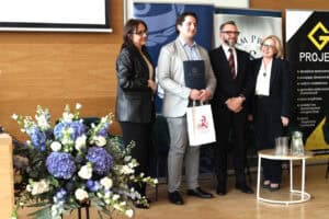 Laureat w szarej marynarce stoi na scenie, trzyma dyplom i białą torbę upominkową. Obok niego stoją trzy uśmiechnięte osoby. W tle widoczny logotyp "Projekt G".
