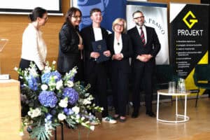 Grupa osób na scenie podczas gratulacji. Młody mężczyzna w czarnej marynarce trzyma dyplom. Obok niego stoją dwie kobiety i mężczyzna w okularach. Wszyscy są uśmiechnięci.
