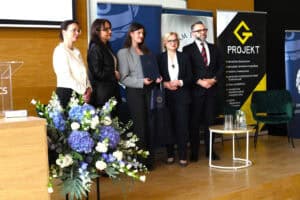 Laureatka w szarej marynarce trzyma dyplom, stoi na scenie w otoczeniu czterech osób. Po lewej stronie widoczny fragment mównicy z napisem UMCS i bukiet niebiesko białych kwiatów.