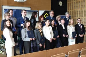 Grupa kilkunastu osób stoi w dwóch rzędach. Wielu laureatów trzyma w dłoniach ciemnoniebieskie dyplomy, statuetki oraz torby upominkowe. W tle widoczny zegar ścienny oraz fragmenty roll-upów.