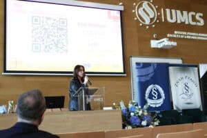 Prelegentka przemawia przy pulpicie. Na dużym ekranie za nią widoczny kod QR oraz link do spotkania online. Z prawej logo Wydziału Prawa i Administracji UMCS.