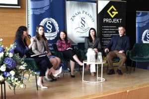 Pięć osób siedzi w ciemnozielonych fotelach na scenie podczas panelu dyskusyjnego. Kobieta po lewej stronie mówi do mikrofonu, trzyma notatnik. W tle widoczne są roll-upy promocyjne Okręgowej Izby Radców Prawnych w Lublinie oraz firmy Projekt G. Na niskim stoliku przed panelistami stoi karafka z wodą i szklanki.