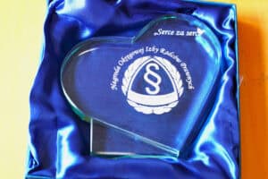 Szklana statuetka w kształcie serca, spoczywająca wewnątrz eleganckiego, ciemnoniebieskiego etui wyściełanego satyną. Na szkle wygrawerowano napis „Serce za serce”, logotyp z paragrafem oraz tekst „Nagroda Okręgowej Izby Radców Prawnych”.