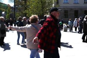 Mężczyzna w czerwono-czarnej koszuli w kratę i czarnej czapce z daszkiem wykonuje zamaszysty gest prawą ręką, w lewej trzyma mikrofon; obok kobieta w ruchu.