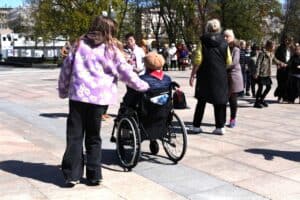 Dziewczyna w jasnofioletowej bluzie w stokrotki kładzie rękę na oparciu wózka inwalidzkiego, na którym siedzi kobieta w brązowym berecie. Wokół nich, na nasłonecznionym bruku, poruszają się inni uczestnicy wydarzenia.