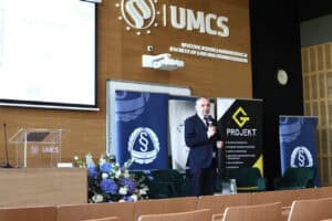 Mężczyzna w garniturze przemawia na scenie auli UMCS. Obok stoją banery Okręgowej Izby Radców Prawnych oraz firmy G-Projekt.