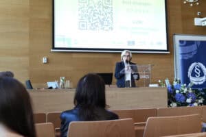 Kobieta w okularach przemawia zza pulpitu w auli uniwersyteckiej. W tle slajd z danymi dostępowymi do transmisji