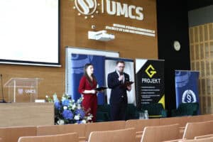 Dwoje młodych prowadzących z teczkami w rękach stoi na scenie obok dekoracji z kwiatów. W tle logotypy uczelni UMCS i partnerów.