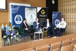 Czworo uczestników panelu dyskusyjnego siedzi w fotelach i rozmawia przez mikrofony. W tle ścianka z logotypami.