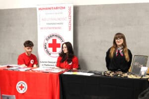 Stoiska informacyjne Polskiego Czerwonego Krzyża oraz Fundacji Głuche Miasto. Wolontariusze siedzą przy stołach z ulotkami i słodyczami.