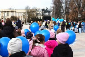 Grupa dzieci i dorosłych idących przez wybrukowany Plac Litewski w Lublinie. Wiele osób niesie jaskrawoniebieskie balony.