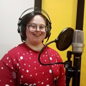 Natalia Opoka stoi w studiu nagraniowym przed mikrofonem, w czerwonym świątecznym swetrze i słuchawkach