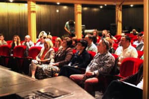 Widok na wypełnioną widownię w teatrze. Publiczność siedzi w czerwonych fotelach, patrzy w stronę sceny. W tle widoczne drewniane kolumny i ciepłe oświetlenie sali.
