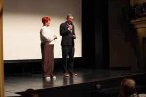Na scenie, stoją od lewej: Maria Pietrusza-Budzyńska oraz Michał Karapuda z mikrofonem w dłoni; w tle duży jasny ekran.