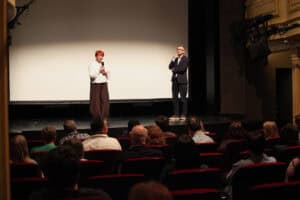 Na scenie teatralnej stoją od lewej: Maria Pietrusza-Budzyńska z mikrofonem oraz Michał Karapuda. Przed nimi siedzą widzowie; widok z widowni.