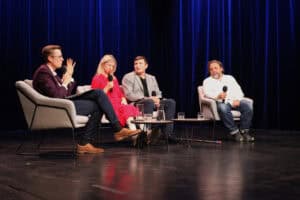 Czworo uczestników panelu dyskusyjnego siedzi na scenie. Na stolikach przed nimi stoją karafki i szklanki z wodą. Uczestnicy gestykulują i trzymają mikrofony