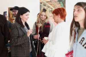 Grupa trzech kobiet rozmawia we wnętrzu galerii; w tle inni uczestnicy wydarzenia