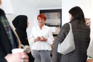 Maria Pietrusza-Budzyńska w białej bluzce uśmiecha się podczas rozmowy z grupą gości w galerii