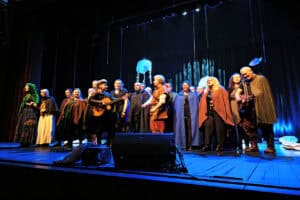 Duża grupa artystów w kostiumach fantasy – pelerynach, tunikach i kapturach w odcieniach brązu, błękitu i zieleni – stoi w szeregu na ciemnej scenie. Nad nimi wiszą ozdobne elementy, w tym świecąca na błękitno konstrukcja przypominająca meduzę. Ciemne tło, niebieskie oświetlenie padające od dołu.