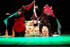 Spektakl "Implication is relation" w wykonaniu Teatru Euforion z Wrocławia. Trójka artystów na ciemnej scenie wykonuje ekspresyjny układ choreograficzny z użyciem kolorowych materiałów w odcieniach czerwieni, błękitu i turkusu. Na środku sceny leży duży rekwizyt w kształcie pająka.