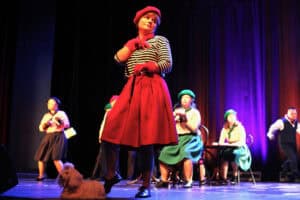 Spektakl "Ponad dachami Paryża" w wykonaniu Zespołu Inscenizacji Teatralnych "Uśmiech" z Kielc. Na pierwszym planie tancerka w czerwonej spódnicy, bluzce w paski i czerwonym berecie trzyma na smyczy pluszowego psa. Za nią, w tle, siedzą inne osoby w podobnych strojach i zielonych beretach.