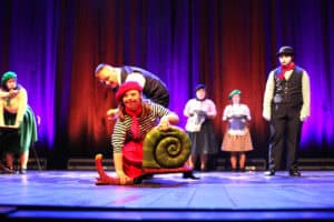 Spektakl "Ponad dachami Paryża" w wykonaniu Zespołu Inscenizacji Teatralnych "Uśmiech" z Kielc. Tancerka w czerwonej spódnicy, bluzce w paski i czerwonym berecie klęczy na podłodze, uśmiecha się do widowni i trzyma ręce na rekwizycie w kształcie dużej, zielonej muszli ślimaka. Nad nią stoi mężczyzna w garniturze. W tle, na fioletowo podświetlonej scenie, siedzą i stoją inne postacie w kostiumach.