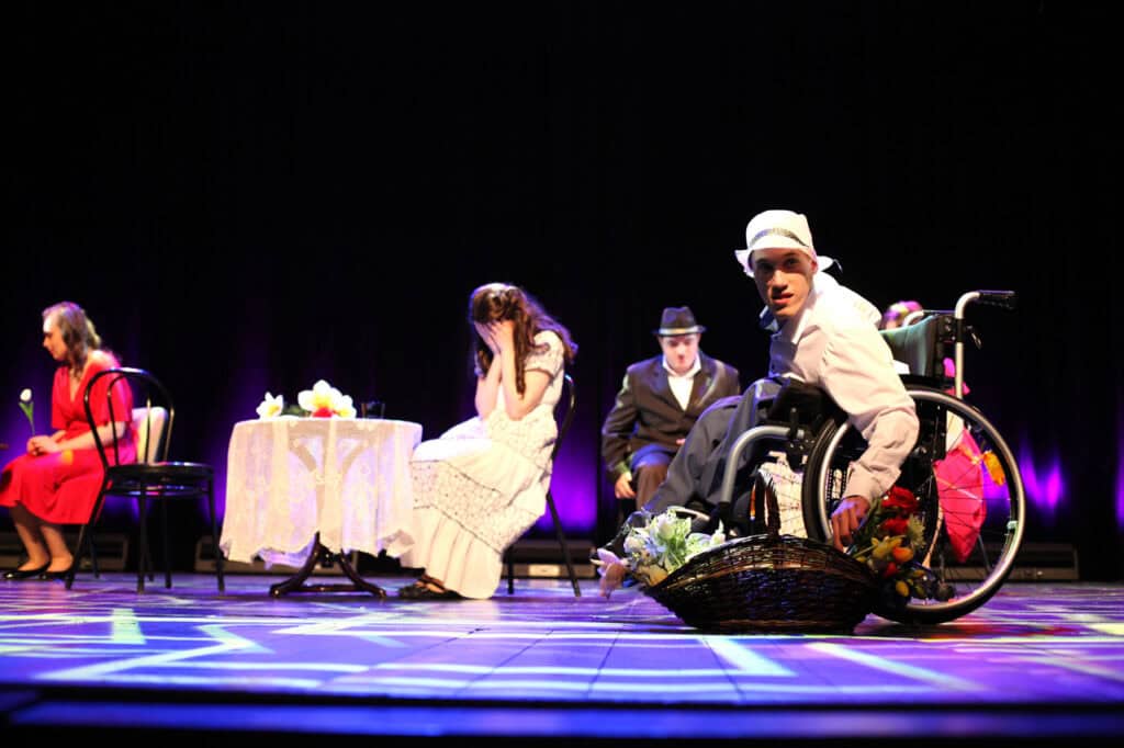 Spektakl Teatru Azumi (Dorohusk) pt. "Lata dwudzieste, lata trzydzieste...". Artysta na wózku inwalidzkim w białym kapeluszu patrzy w stronę widowni, trzyma koszyk z kwiatami. W tle przy stolikach siedzą inni artyści.