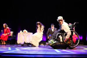 Spektakl Teatru Azumi (Dorohusk) pt. "Lata dwudzieste, lata trzydzieste...". Artysta na wózku inwalidzkim w białym kapeluszu patrzy w stronę widowni, trzyma koszyk z kwiatami. W tle przy stolikach siedzą inni artyści.