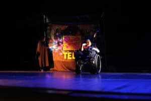 Spektakl pt. "Czarmedia" W wykonaniu Teatru Tacy Sami z Matczyna. Ciemna scena z teatralnym oświetleniem. Pośrodku siedzi aktor na wózku inwalidzkim. Za nim widać tło z kolorowymi projekcjami świetlnymi i fragmentem napisu „TEL”. Po lewej i prawej stronie, w cieniu, stoją dwie postacie w pelerynach z kapturami.
