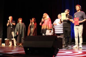 Na scenie stoi grupa osób z Teatru Euforion; w centrum kobieta w różowo-pomarańczowej tunice mówi do mikrofonu, trzymając kartkę. Po prawej stronie dwaj mężczyźni dumnie prezentują swoje dyplomy – jeden żółty, drugi czerwony. Wszyscy są oświetleni reflektorami na ciemnym tle