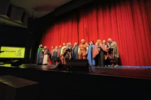 Artyści w kostiumach fantasy stoją w rzędzie na ciemnej scenie na tle dużej, zaciągniętej czerwonej kurtyny. Po lewej stronie widać ekran projekcyjny z wyświetlonym napisem „FANTASY” oraz grafiką. Grupa ubrana jest w peleryny, kaptury i stroje nawiązujące do średniowiecza