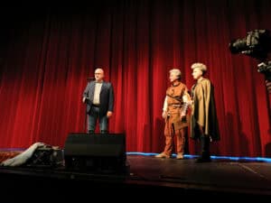 Na scenie na tle czerwonej kurtyny stoją trzy osoby. Po lewej Janusz Iwanicki w garniturze mówi do mikrofonu. Obok niego Michał Stanowski w brązowej tunice i Karolina Czajka w złotej pelerynie.