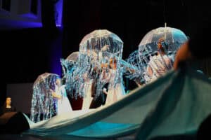 Grupa dziecięca w białych sukienkach wykonuje układ taneczny na scenie. Tancerki trzymają przezroczyste parasole, z których zwisają błyszczące, srebrne i niebieskie paski. Na pierwszym planie uniesiony kawałek jasnoniebieskiego materiału, tworzącego efekt fali.