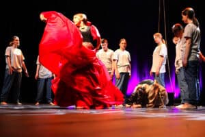 Spektakl "W cieniu katedry" Teatru Mlekołaki z Olbięcina. W centrum młoda tancerka w czerwonej, rozłożystej spódnicy i czarnym topie wykonuje ekspresyjny ruch, unosząc materiał spódnicy. Otaczają ją inne osoby w szarych t-shirtach i dżinsach, stojące nieruchomo