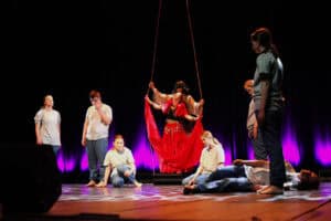 Spektakl "W cieniu katedry" Teatru Mlekołaki z Olbięcina. Scena teatralna z fioletowo-różowym podświetleniem. Na podwyższeniu, tancerka i aktor trzymają długie liny. Wokół nich na podłodze siedzą i leżą inne osoby w szarych t-shirtach i dżinsach.