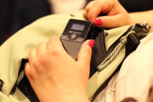 Kobiece dłonie trzymają małe, czarne urządzenie elektroniczne marki Retekess, służące do audiodeskrypcji lub tłumaczenia symultanicznego.