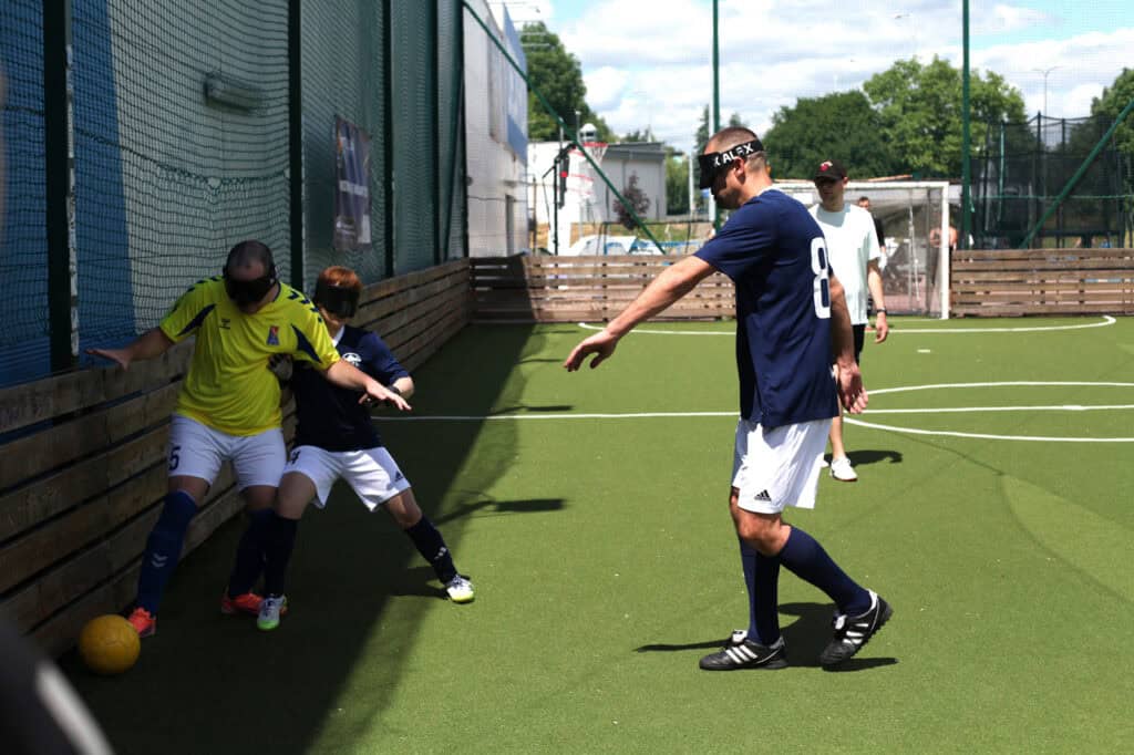 Dwoje zawodników blind footballu w opaskach sensorycznych walczy o piłkę przy drewnianej bandzie boiska. Z boku nadchodzi trzeci gracz z rozłożonymi rękami, lokalizując dźwięk piłki.