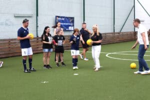 Uczestnicy i organizatorzy stoją na środku boiska trawiastego. Anna Augustyniak trzyma żółtą piłkę do blind footballu, a obok stoją zawodnicy w strojach sportowych oraz wolontariusze.