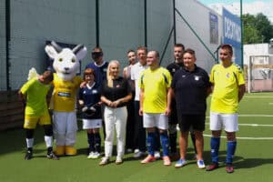 Pamiątkowe zdjęcie grupowe uśmiechniętych uczestników, piłkarzy blind footballu oraz organizatorów na boisku. Po lewej stronie stoi biała maskotka Koziołka w żółtej koszulce z napisem Lublin.