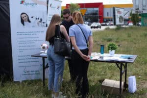 Dwie kobiety przy plenerowym stoisku kliniki okulistycznej Optegra, rozmawiają z pracownikiem. Na stole leżą ulotki i stoi model ludzkiego oka, a obok znajduje się baner reklamowy z ofertą laserowej korekcji wzroku.
