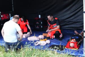 Ratownicy medyczni w pomarańczowych strojach prowadzą pokaz pierwszej pomocy w namiocie. Na niebieskiej plandece leżą fantomy szkoleniowe niemowlęcia i osób dorosłych, defibrylator AED oraz nosze typu deska, a z boku przygląda się im klęczący mężczyzna.
