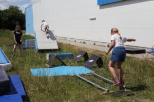 Dziewczynka w jeansowych spodenkach ćwiczy równowagę na metalowych rurkach zamontowanych na trawie. W tle inna dziewczynka wspina się na skośną przeszkodę, a całości przygląda się stojąca z boku kobieta.