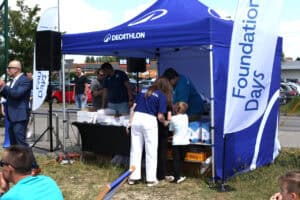 Pracownicy i uczestnicy wydarzenia zgromadzeni wokół niebieskiego namiotu z napisami Decathlon oraz Foundation Days. Na stole pod namiotem przygotowano materiały w białych kartonach oraz kartony z wodą.