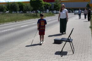 Chłopiec w opasce zasłaniającej oczy i koszulce piłkarskiej idzie po chodniku, używając białej laski. Kilka kroków przed nim stoi dorosła kobieta w okularach przeciwsłonecznych, a na drodze chłopca ustawiono przeszkody w postaci krzesła i pudełka.