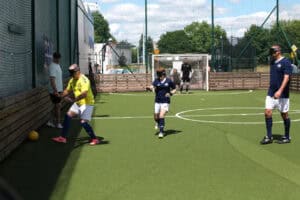 Mecz blind footballu na ogrodzonym miniboisku ze sztuczną trawą. Trzej zawodnicy w opaskach sensorycznych na oczach lokalizują żółtą piłkę, a w tle, w bramce, stoi bramkarz.